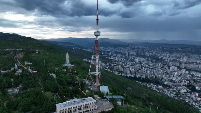 Aerial view of the Funicular Restaurant at Mtatsminda Park during sunset. Dynamic drone shot of a TV tower with Tbilisi city in the background.