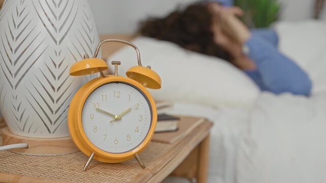 Woman rubbing her eyes beside a yellow alarm clock on a rattan bedside table with books in a building bedroom; sleepy morning.