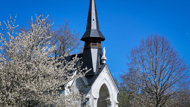 Aerial view of Chapelle Notre-Dame de Riantmont featuring its dark spire and white stone facade surrounded by blossoming spring trees under a clear blue sky Vesancy, Auvergne-Rhone-Alpes, France.