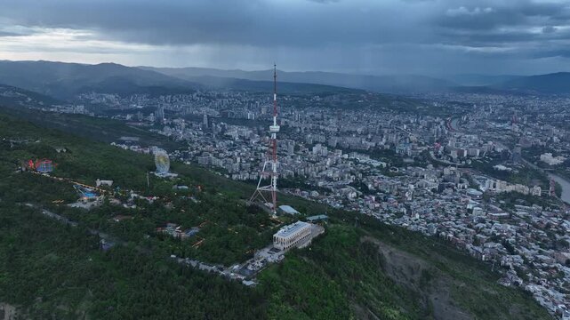 Aerial view of the Funicular Restaurant at Mtatsminda Park during sunset. Dynamic drone shot of a TV tower with Tbilisi city in the background.
