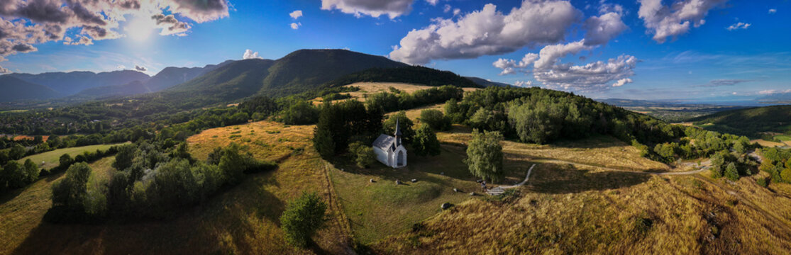 Aerial view of Chapelle Notre-Dame de Riantmont, a small white chapel on a grassy hill surrounded by lush green trees and rolling mountains under a blue sky in Vesancy, Auvergne-Rhone-Alpes, France.