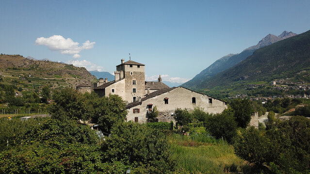 Aerial view of a medieval castle surrounded by lush green trees and vineyards with majestic mountains in the background under a clear blue sky Aosta Valley, Aosta Valley, Italy.