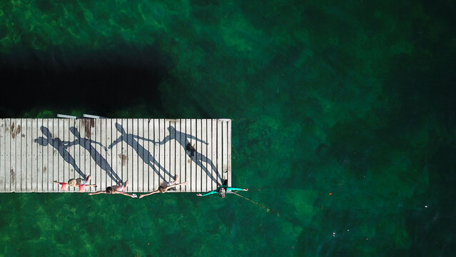 Aerial view of four children standing on a wooden pier with arms outstretched casting long shadows over turquoise water in Saint-Prex, Vaud, Switzerland.
