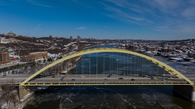 Aerial view of the yellow Daniel Carter Beard Bridge spanning the icy Ohio River with snowy banks and city buildings under a clear blue sky Cincinnati, Ohio, United States.