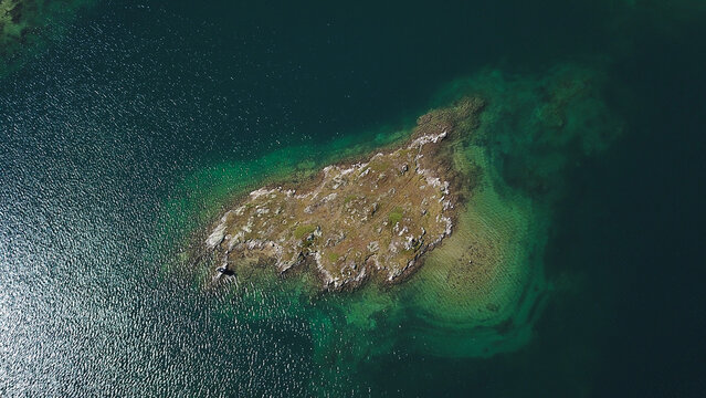 Aerial view of a small rocky island surrounded by turquoise alpine water and sunlight reflections in Obergoms, Valais, Switzerland.