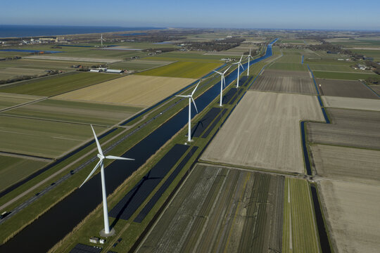 Aerial view of a row of modern wind turbines and solar panels along a straight canal through flat agricultural fields under a clear blue sky Netherlands.
