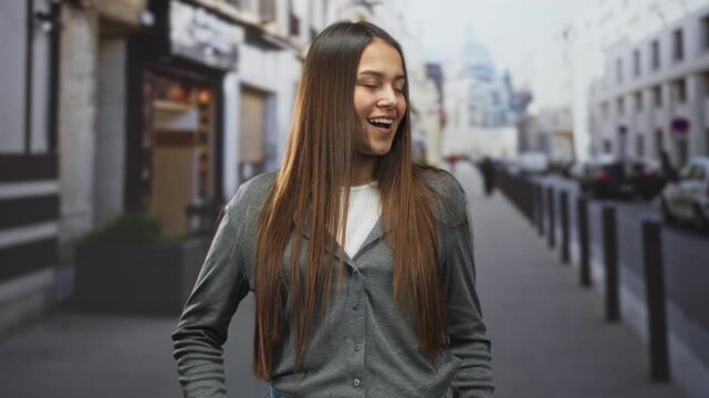 Woman with mouth open in a gasp, long hair over a gray cardigan, hands near pockets, standing on a street with bollards and storefronts; youthful curiosity surprise.