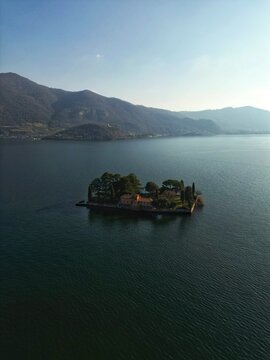 Aerial view of Isola di Loreto with its neo-Gothic villa and lush gardens on Lake Iseo surrounded by calm water and distant mountains Monte Isola, Lombardy, Italy.