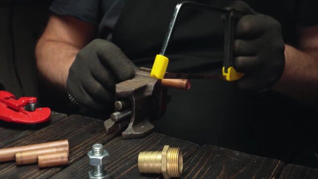 Skilled technician using a hacksaw to cut a copper pipe secured in a vise on a wooden workbench surrounded by various plumbing components
