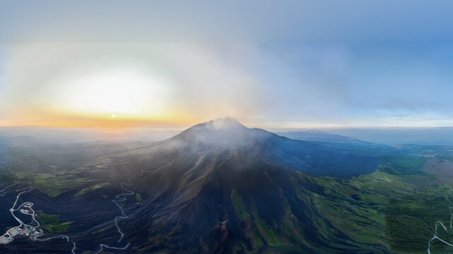 Aerial view of Mount Etna with steam rising from the crater and winding roads on the volcanic slopes at sunset Castiglione di Sicilia, Sicily, Italy.