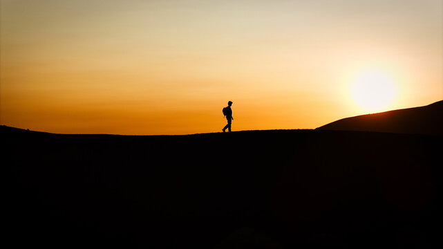 Aerial view of a lone hiker silhouetted against the setting sun on the volcanic slopes of Mount Etna under a warm orange sky in Castiglione di Sicilia, Sicily, Italy.