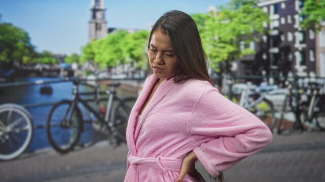 Woman in pink bathrobe holds lower back with a pained face beside a canal and bicycles near a city street bridge; pain backache.
