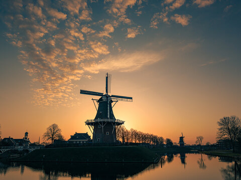 Aerial view of a traditional Dutch windmill silhouette against a golden sunset sky with scattered clouds and water reflections in Dokkum, Friesland, Netherlands.