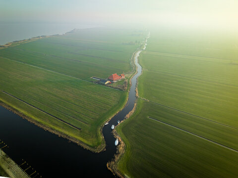 Aerial view of a narrow winding canal through green polders with a farmhouse and small boats under a hazy sky Sloten, Friesland, Netherlands.