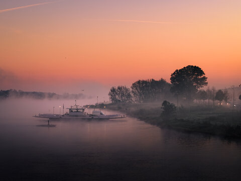 Aerial view of a ferry boat crossing the misty Meuse river during a vibrant orange sunrise with thick fog over the water and trees on the bank in Kessel, Limburg, Netherlands.