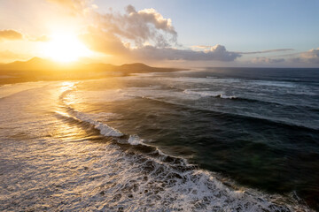 Obraz premium People walk along Famara beach in Lanzarote during a sunset. Waves crash on the shore while the sun sets behind distant mountains