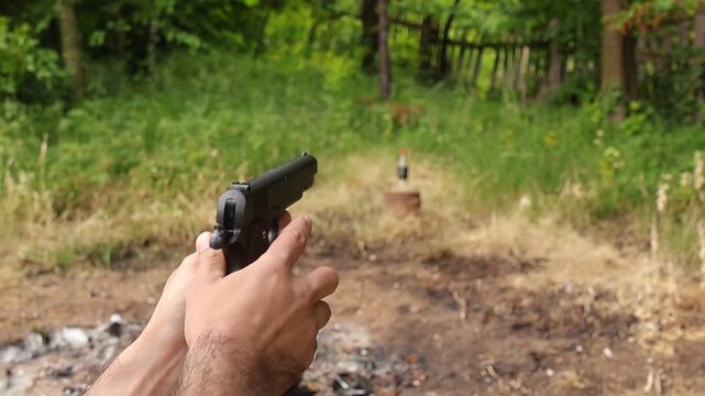 Close up of a person holding a handgun with both hands while aiming at a bottle outdoors in a rural area, focused shooting practice scene with blurred natural background in daylight.