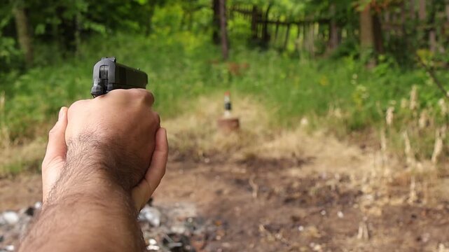 Close up shot of hands firing an airsoft pistol at a bottle target in a forest clearing, capturing the moment the bottle falls after a successful hit during shooting practice.