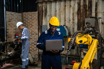 Engineer in yellow hard hat holds laptop. Yellow robotic arm operates nearby. Another worker uses handheld device. Brick walls and machinery surround the scene. Industrial workshop setting © VStudio