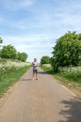 Obraz premium Man standing in distance on empty countryside road in green landscape, peaceful solitude, outdoor travel, summer exploration, rural freedom.