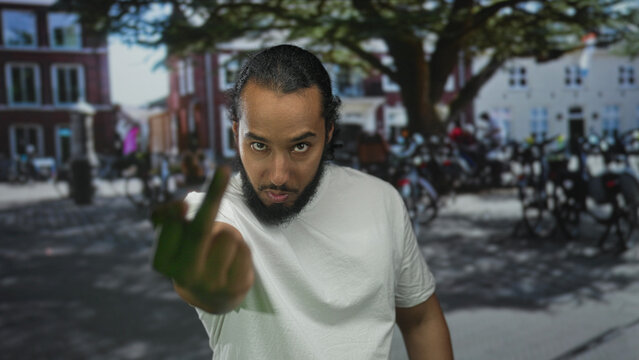 Man shows middle finger in street near parked motorcycles and tree by brick buildings, wearing white shirt and beard; defiance.