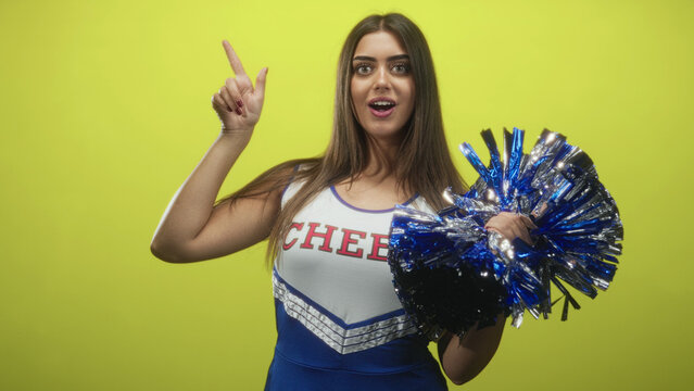 Woman points finger up, mouth open, holding metallic blue pompom, wearing cheer uniform with cheer lettering in studio; team spirit enthusiasm.