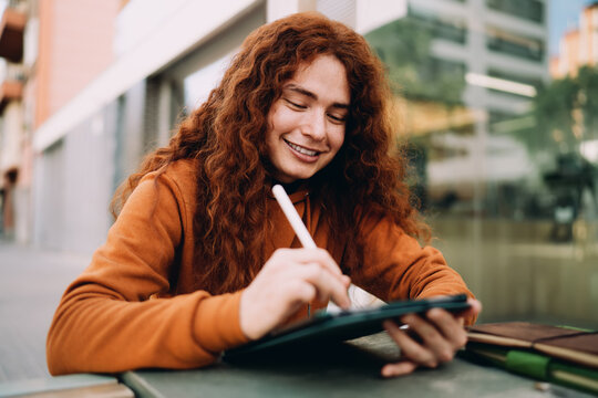 Close up of cheerful multiracial model with long curly ginger hair using stylus for digital sketching on tablet. Creative professional working on web design project, mobile application interface.