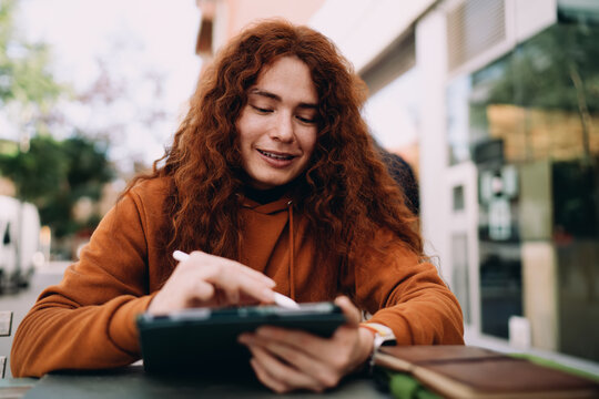 Young multiracial model with curly hair focusing on digital tablet screen using stylus. Software development, graphic design, remote education process, student working on project in urban cafe.