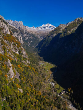 Aerial view of Val di Mello valley with autumn foliage on steep granite cliffs and snow-capped peaks under a clear blue sky Val Masino, Lombardia, Italy.