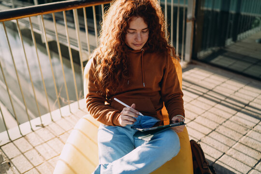 Top view of focused red-haired professional using stylus for digital sketching on tablet outdoors. Creative illustrator working on project, remote freelance job, web design process, education.
