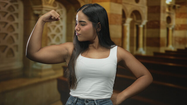 Woman flexes biceps and clenches fists while smiling, wearing white tank top and jeans, in ornate stone building hall with arched columns; confidence empowerment.