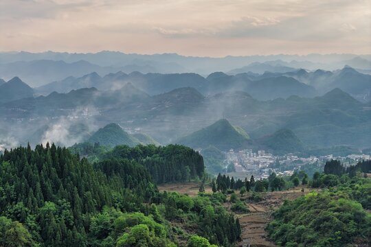 Aerial view of misty karst mountains and a traditional village nestled among lush green forests under a soft overcast sky Gaoxing Miao Village, Guizhou, China.
