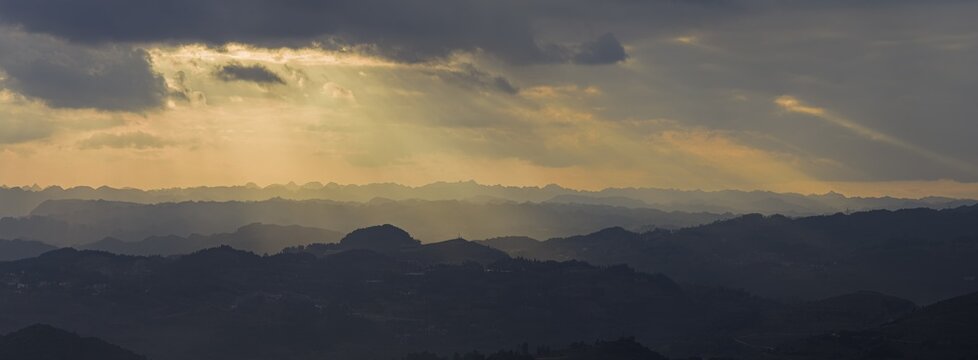 Aerial view of rolling mountain ridges and silhouetted hills under a dramatic sunset sky with golden sunbeams breaking through dark clouds in Gaoxing Miao Village, Guizhou, China.