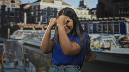 Fototapeta premium Young woman with fists raised in a boxing pose wearing a blue shirt and denim jeans in front of a city building; playful confidence.