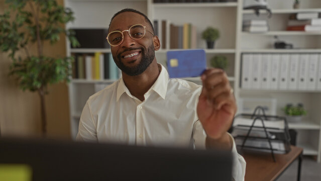 Man holding creditcard with hand at office desk near laptop monitor wearing glasses and braces; financial confidence.