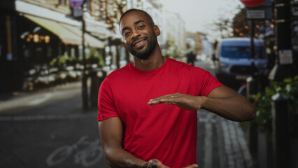 Obraz premium Man in red t shirt making a measuring gesture with hands on an urban street near cafe seating; casual conversation friendly.