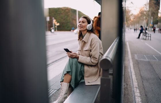 Caucasian woman sitting at city bus stop waiting for transport, female traveler with headphones and smartphone looking away, urban environment, authentic representation, real people life.