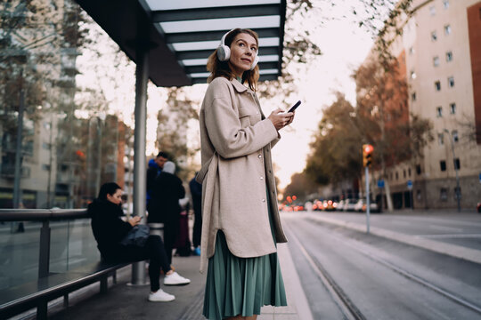 Caucasian woman waiting at urban bus stop with headphones, stylish lady using smartphone app for navigation, city transport connectivity, digital nomad travel, authentic representation.
