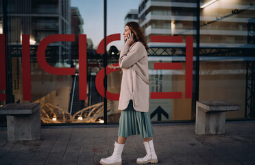 Fototapeta premium Mature woman walking city street talking mobile phone, autumn casual outfit, trendy pleated skirt, white boots, outdoor communication, 5G wireless connection, cellular network, urban lifestyle.