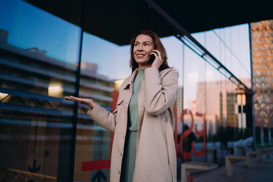 Middle aged business woman using mobile phone for international call, standing outdoors, urban glass architecture, evening light, wireless communication, technology, holding gadget, talking.