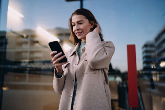 Middle aged woman browsing smartphone, reading notification or text message, standing near office building, evening urban lighting, mobile application usage, wireless data, digital nomad concept.