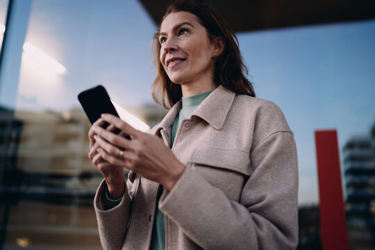 Confident lady looking away while holding smartphone, waiting for message or call, standing in urban environment, evening light, wireless connectivity, digital lifestyle, career growth concept.