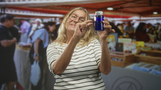 Woman holding blue pill bottle, pinching fingers near face to show tiny dose in indoor market building; wellness reassurance contentment.