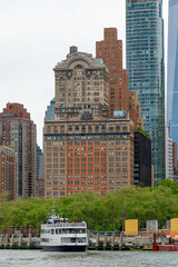 Fototapeta premium Historic Whitehall building and ferry boat at Manhattan pier. Old architecture skyscraper in Lower Manhattan with passenger ferry at the dock and green trees in foreground.