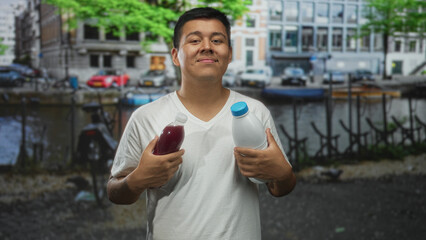 Fototapeta premium Young man holding two bottles with hands at chest on street by canal and building, wearing white t shirt; cheerful confidence.