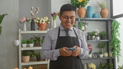 Fototapeta premium Man florist in apron holds smartphone and smiles while texting in a building florist shop surrounded by potted flowers and vases; small business content.