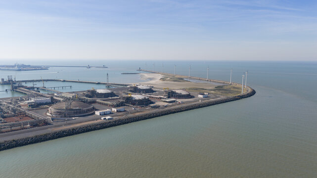 Aerial view of Fluxys LNG Terminal Zeebrugge featuring large storage tanks and a line of wind turbines along the breakwater under a clear blue sky in Brugge, Vlaanderen, Belgium.