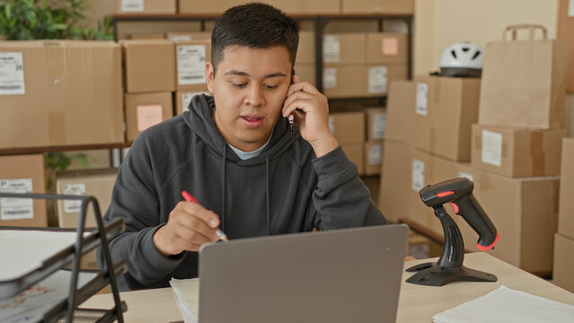 Man holds smartphone to ear and writes by laptop amid stacked parcel boxes and barcode scanner in a building; small business stress.