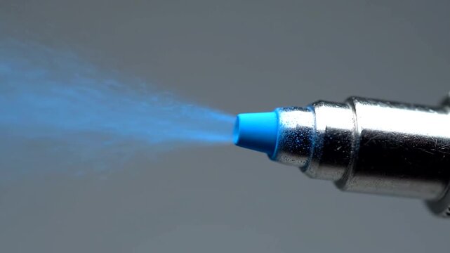 Close-up of a fuel injector spraying a fine mist of blue liquid against a gray background.