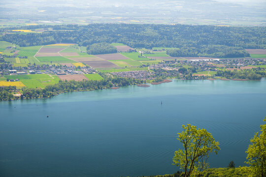 Elevated spring panorama over Lake Biel with shoreline villages, farmland mosaics, and forested hills in the Swiss Jura.